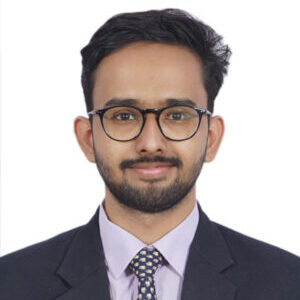 Image of Kartik wearing spectacles, dressed in a black blazer, purple shirt and a checkered tie with a smirk for the headshot